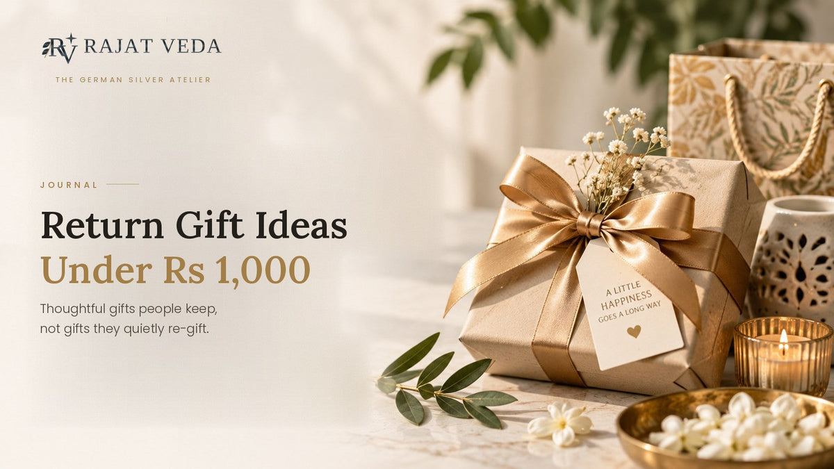 A return gift wrapped in kraft paper and gold ribbon with a hand-written tag, beside a gift bag, a lit candle and jasmine flowers