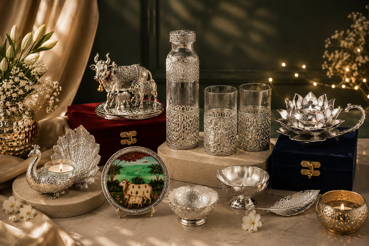 German Silver serveware and pooja pieces on a marble table — a Kamdhenu cow idol, jali water bottle and tumblers, a Pichwai plate, lotus diya and peacock tealight holder